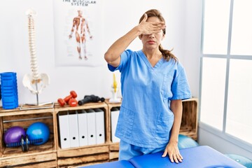 Middle age hispanic physiotherapist woman working at pain recovery clinic covering eyes with hand, looking serious and sad. sightless, hiding and rejection concept