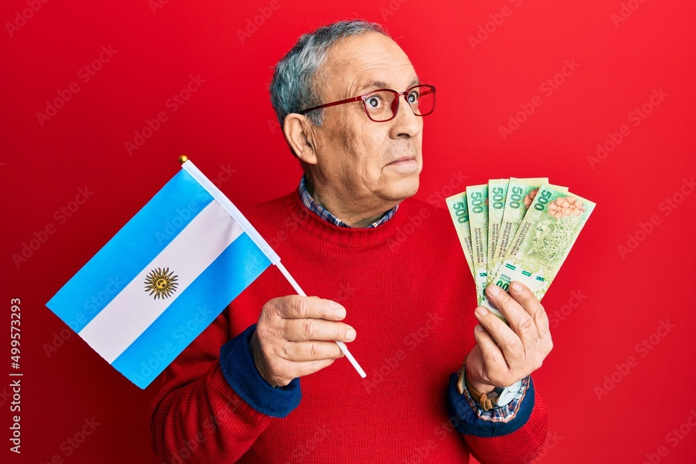 Wall mural Handsome senior man with grey hair holding argentine flag and pesos banknotes smiling looking to the side and staring away thinking.