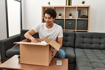 Young hispanic man unboxing cardboard box sitting on the sofa at home.