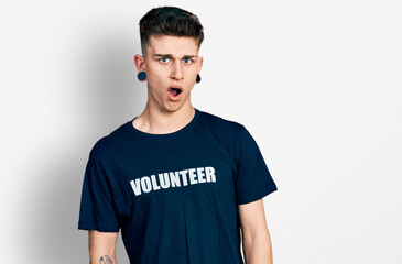 Young caucasian boy with ears dilation wearing volunteer t shirt in shock face, looking skeptical and sarcastic, surprised with open mouth