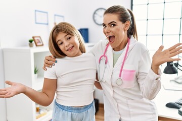 Pediatrician woman working at the clinic with little girl celebrating achievement with happy smile and winner expression with raised hand