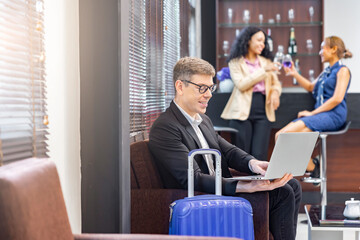 Businessman reading or writing email on laptop, relaxing laughing watching funny movie on laptop, sitting with suitcase at the airport lounge Blurred background of passengers at counter serving drink.