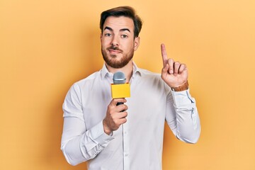 Handsome caucasian man with beard holding reporter microphone surprised with an idea or question pointing finger with happy face, number one - obrazy, fototapety, plakaty