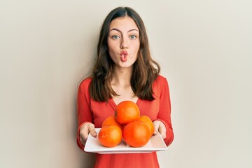 Young brunette woman holding plate with fresh oranges making fish face with mouth and squinting eyes, crazy and comical.