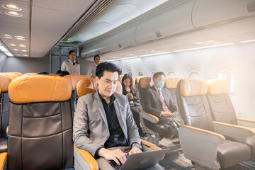Successful entrepreneur smiling in satisfaction as he checks information on his laptop computer while sitting comfortably in the aircraft. Cabin of airplane with passengers on background.