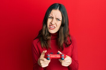 Young brunette woman playing video game holding controller clueless and confused expression. doubt...