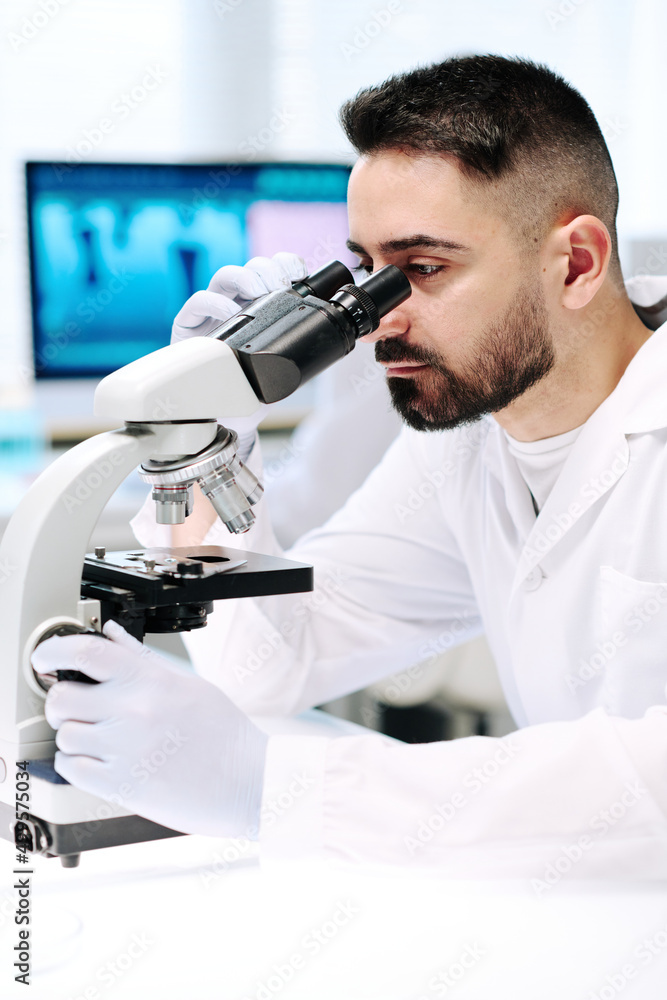 Poster young bearded man in lab coat and gloves looking at bacteria in microscope while sitting by his work
