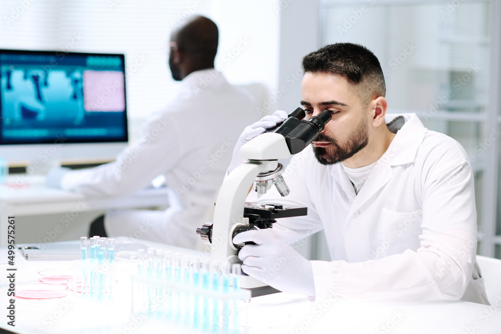 Poster young serious male chemist in whitecoat and gloves looking in microscope by desk with group of flask