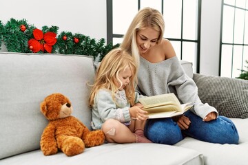 Mother and daughter reading book sitting by christmas decor at home