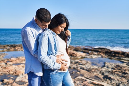 Young latin couple expecting baby touching belly at seaside
