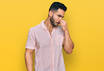 Hispanic man with beard wearing casual shirt smelling something stinky and disgusting, intolerable smell, holding breath with fingers on nose. bad smell