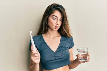 Young caucasian woman holding electric toothbrush and water glass making fish face with mouth and squinting eyes, crazy and comical.