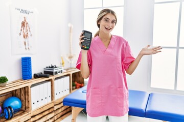 Young physiotherapist woman working at pain recovery clinic holding dataphone celebrating achievement with happy smile and winner expression with raised hand
