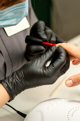 Manicure varnish painting. Close-up of a manicure master wearing rubber black gloves applying red varnish on a female fingernail in the beauty salon