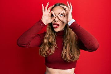Young blonde woman wearing turtleneck sweater doing ok gesture like binoculars sticking tongue out,...
