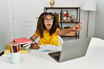 Young african american girl doing homework at home pointing with finger surprised ahead, open mouth amazed expression, something on the front