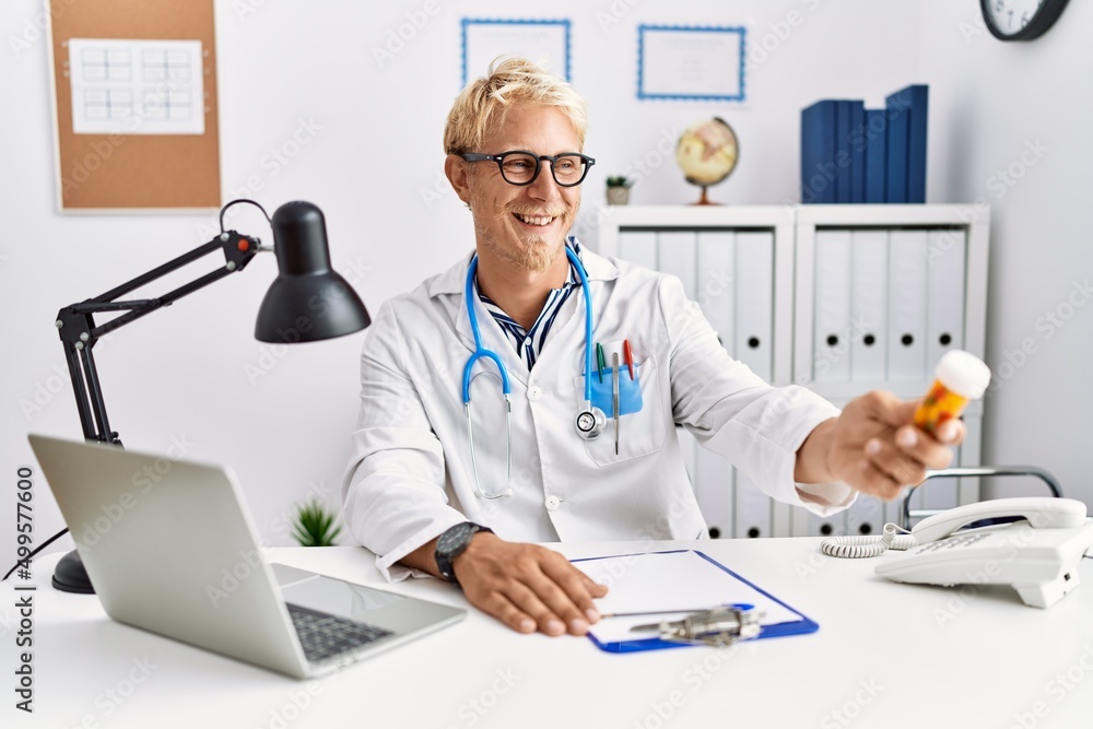 Sticker Young caucasian man wearing doctor uniform holding pills at clinic