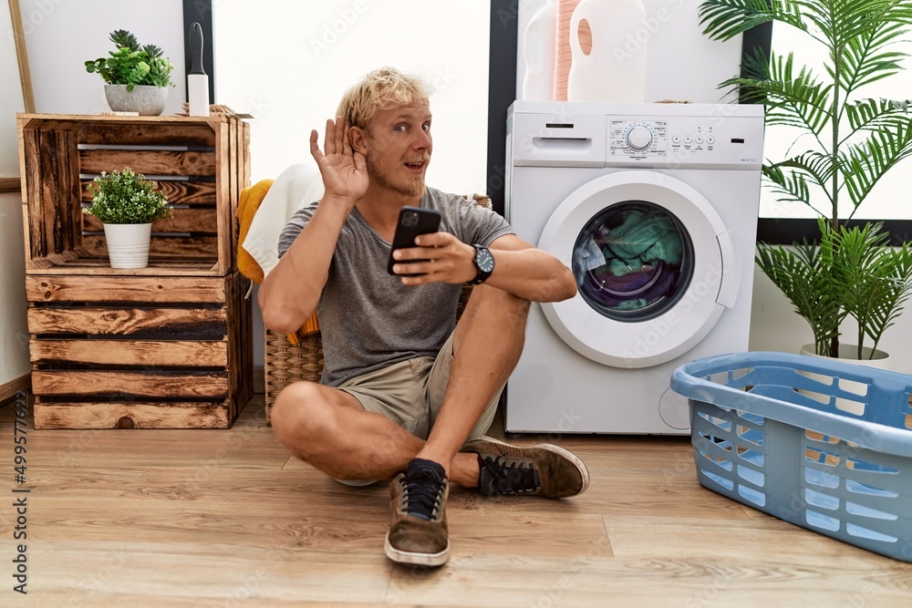 Sticker Young blond man doing laundry using smartphone smiling with hand over ear listening an hearing to rumor or gossip. deafness concept.