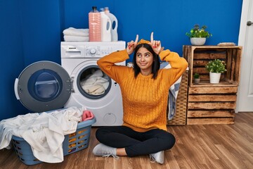 Young hispanic woman doing laundry doing funny gesture with finger over head as bull horns