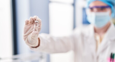 Young blonde woman wearing scientist uniform and medical mask holding covid-19 vaccine dose at laboratory