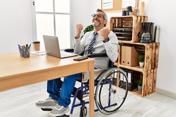 Middle age hispanic man working at the office sitting on wheelchair very happy and excited doing winner gesture with arms raised, smiling and screaming for success. celebration concept.
