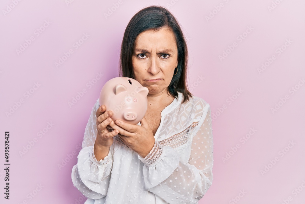 Wall mural middle age hispanic woman holding piggy bank skeptic and nervous, frowning upset because of problem.