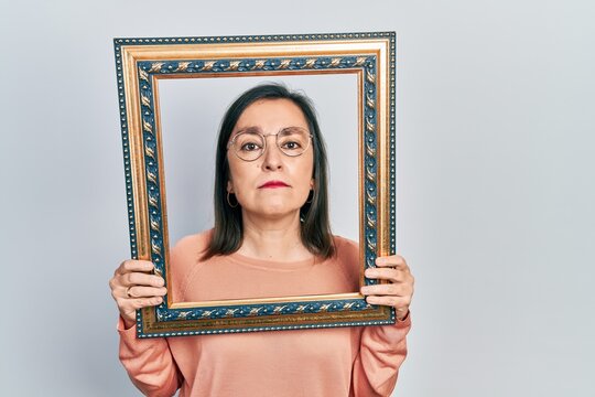 Middle age hispanic woman holding empty frame relaxed with serious expression on face. simple and natural looking at the camera.