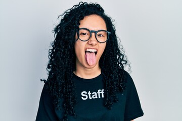 Young hispanic woman with curly hair wearing staff t shirt sticking tongue out happy with funny...