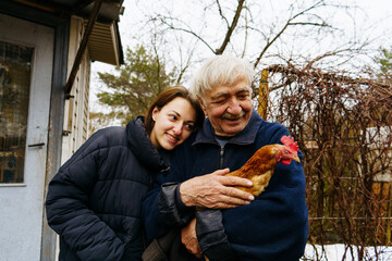 a gray-haired elderly man farmer holds a brown chicken in his hands. his 25-year-old adult granddaughter is standing next to him and smiling