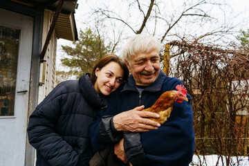 Grandfather and granddaughter are holding a chicken in their hands, looking at it and hugging standing on the farm
