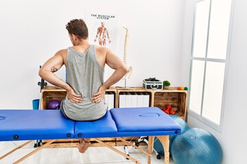 Young hispanic man touching injury back sitting on massage board at clinic