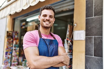 Fototapeta premium Young hispanic worker wearing apron standing with arms crossed gesture at newspaper store.