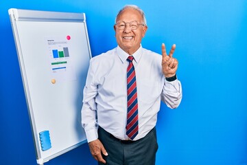 Senior man with grey hair standing by business blackboard smiling with happy face winking at the camera doing victory sign with fingers. number two.