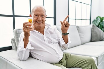 Fototapeta premium Senior man holding pills cheerful with a smile on face pointing with hand and finger up to the side with happy and natural expression