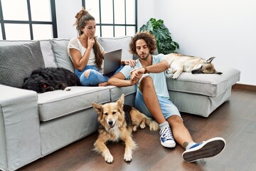 Young hispanic couple with dogs relaxing at home checking the time on wrist watch, relaxed and...