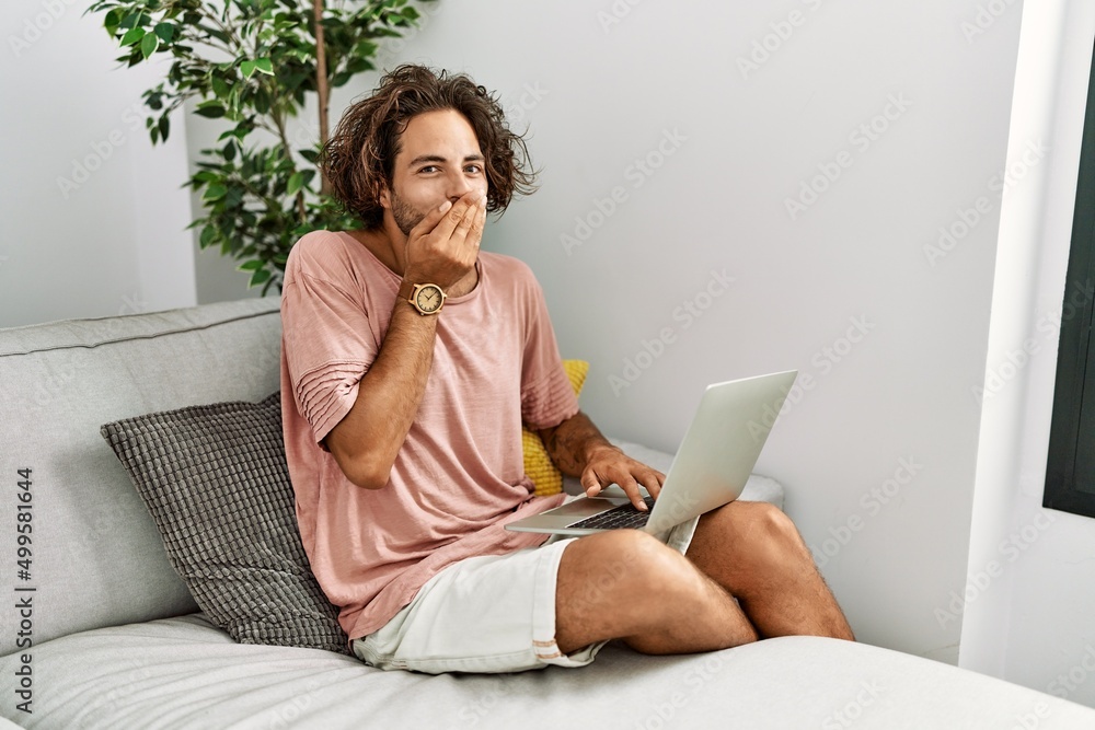 Sticker young hispanic man sitting on the sofa at home using laptop laughing and embarrassed giggle covering