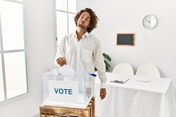 Young hispanic man voting putting envelop in ballot box making fish face with lips, crazy and...
