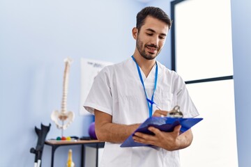 Young hispanic man wearing physiotherapist uniform writing on clipboard at rehab clinic