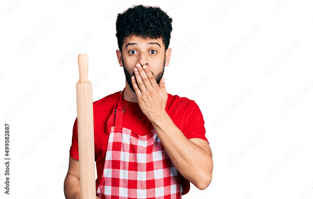 Wall mural Young arab man with beard wearing professional baker apron holding kneading roll covering mouth with hand, shocked and afraid for mistake. surprised expression