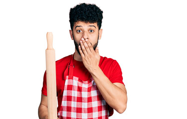 Young arab man with beard wearing professional baker apron holding kneading roll covering mouth...