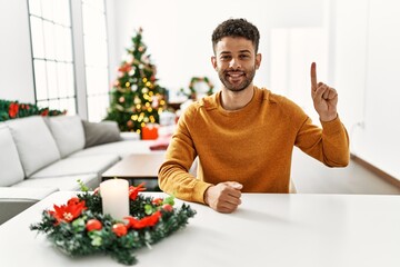 Arab young man sitting on the table by christmas tree showing and pointing up with finger number one while smiling confident and happy.