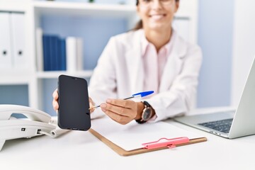 Young hispanic woman wearing doctor uniform showing smartphone screen at clinic