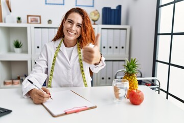 Young redhead woman nutritionist doctor at the clinic approving doing positive gesture with hand,...