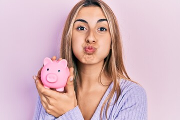 Beautiful hispanic woman holding piggy bank puffing cheeks with funny face. mouth inflated with...