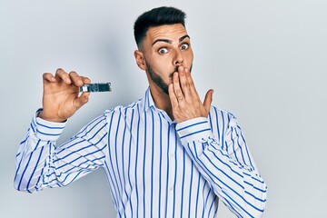 Young hispanic man with beard holding ssd memory covering mouth with hand, shocked and afraid for...