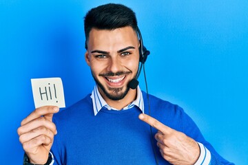 Young hispanic man with beard wearing operator headset showing hi greeting smiling happy pointing...