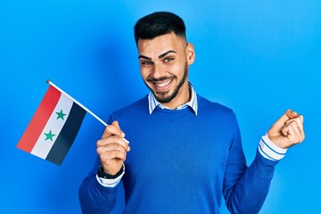 Young hispanic man with beard holding syria flag screaming proud, celebrating victory and success very excited with raised arm