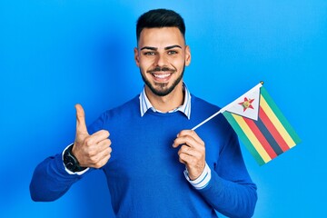 Young hispanic man with beard holding zimbabwe flag smiling happy and positive, thumb up doing excellent and approval sign