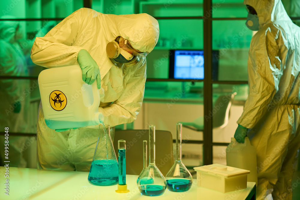 Poster one of three scientists in protective coveralls and respirators pouring blue liquid in big test tube