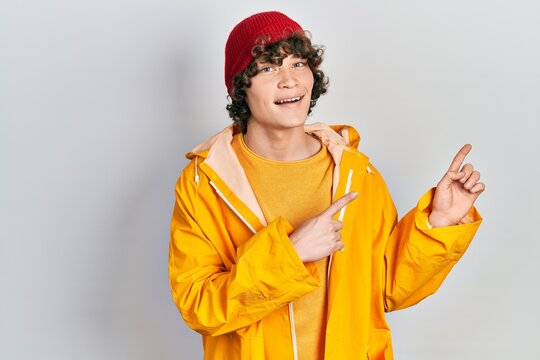 Handsome young man wearing yellow raincoat smiling and looking at the camera pointing with two hands and fingers to the side.